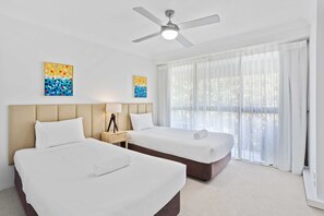 Habitación de hotel con dos camas, ventilador de techo, obras de arte en las paredes y un amplio ventanal con cortinas.