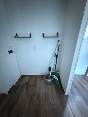 A room with a wooden floor, a broom leaning against the wall, and a shelf with a small item on it. - Lovely Per Diem (Angleton)