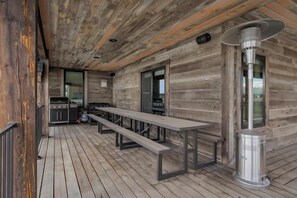 Una terraza de madera con un calentador metálico, un banco largo y una cocina visible a través de una puerta abierta.
