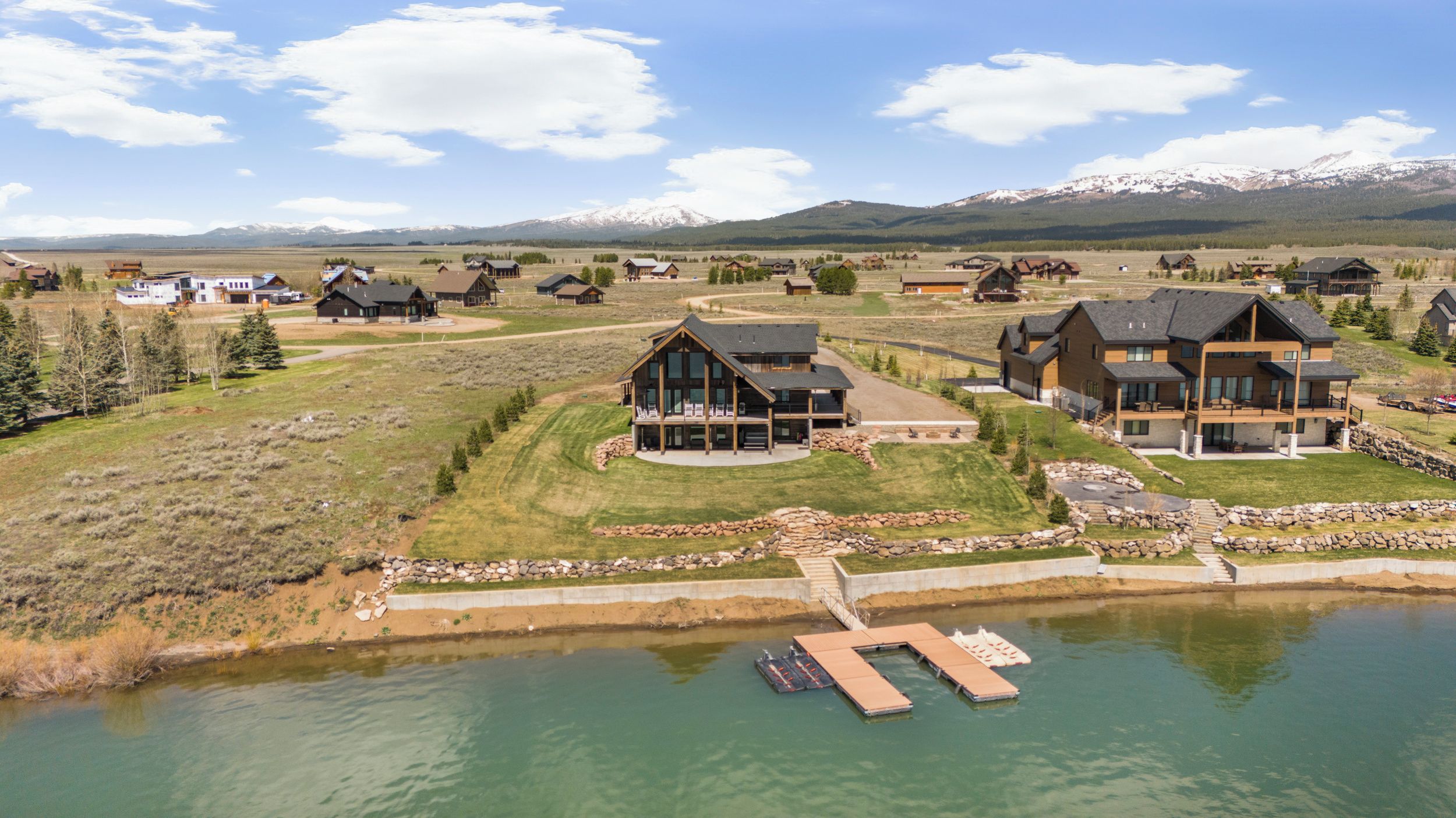 A large, modern house with a dock and a boat, surrounded by a lake and mountains in the background.