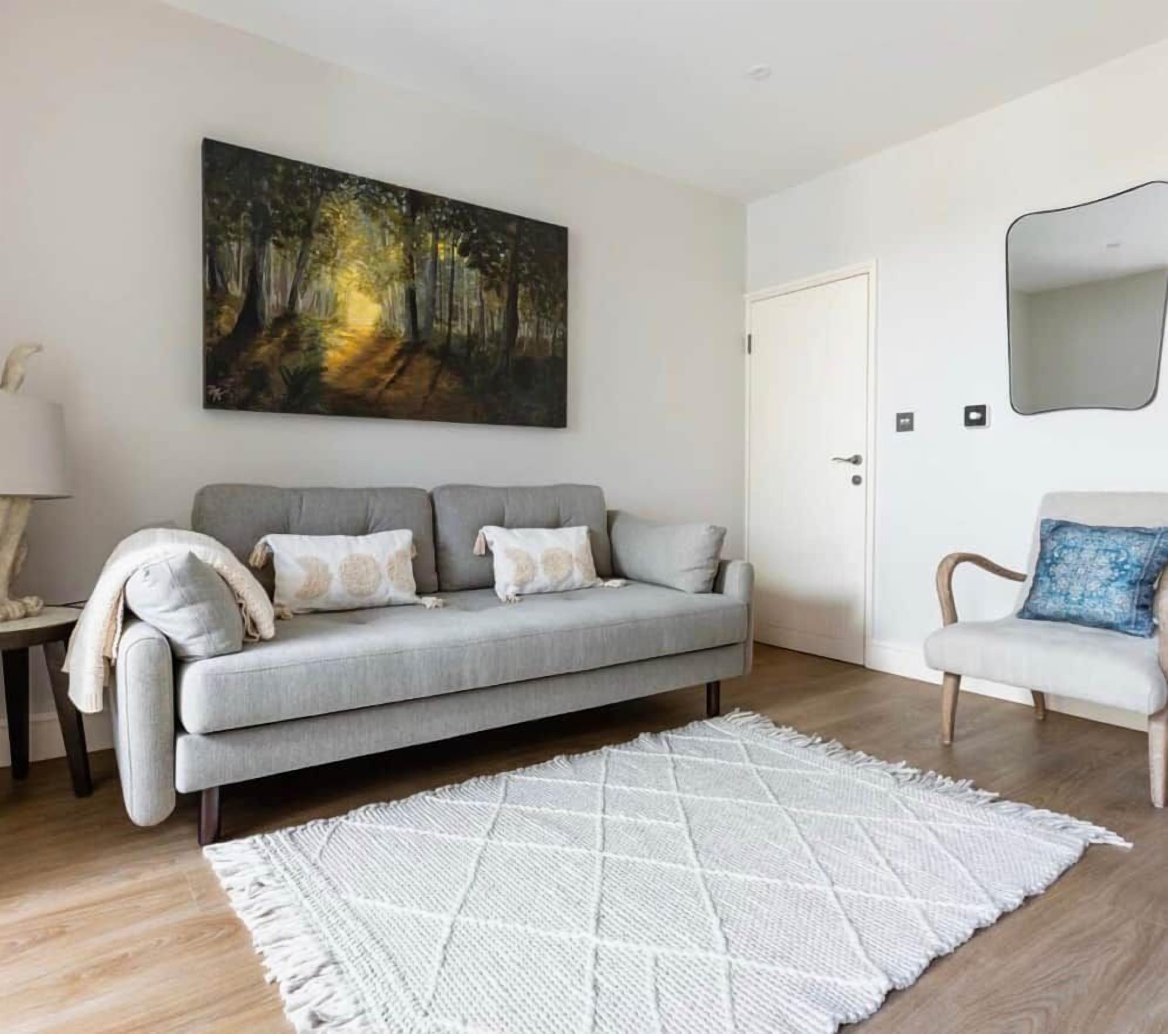 Ein Wohnzimmer mit einem grauen Sofa, einem weißen Teppich und einem Gemälde von einem Wald.
