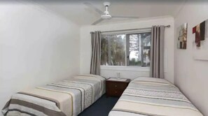 Un petit logement composé d’une seule pièce, comprenant deux lits, un ventilateur de plafond, une fenêtre avec des rideaux et une petite table de chevet.