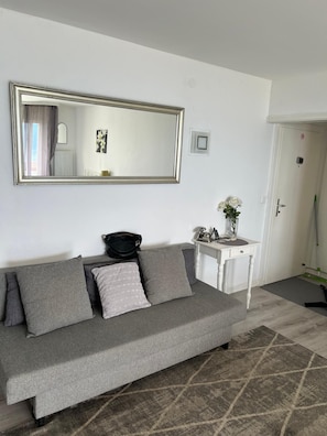 A neutral-colored sofa with cushions, a white side table with a vase and a lamp, and a large mirror on the wall.