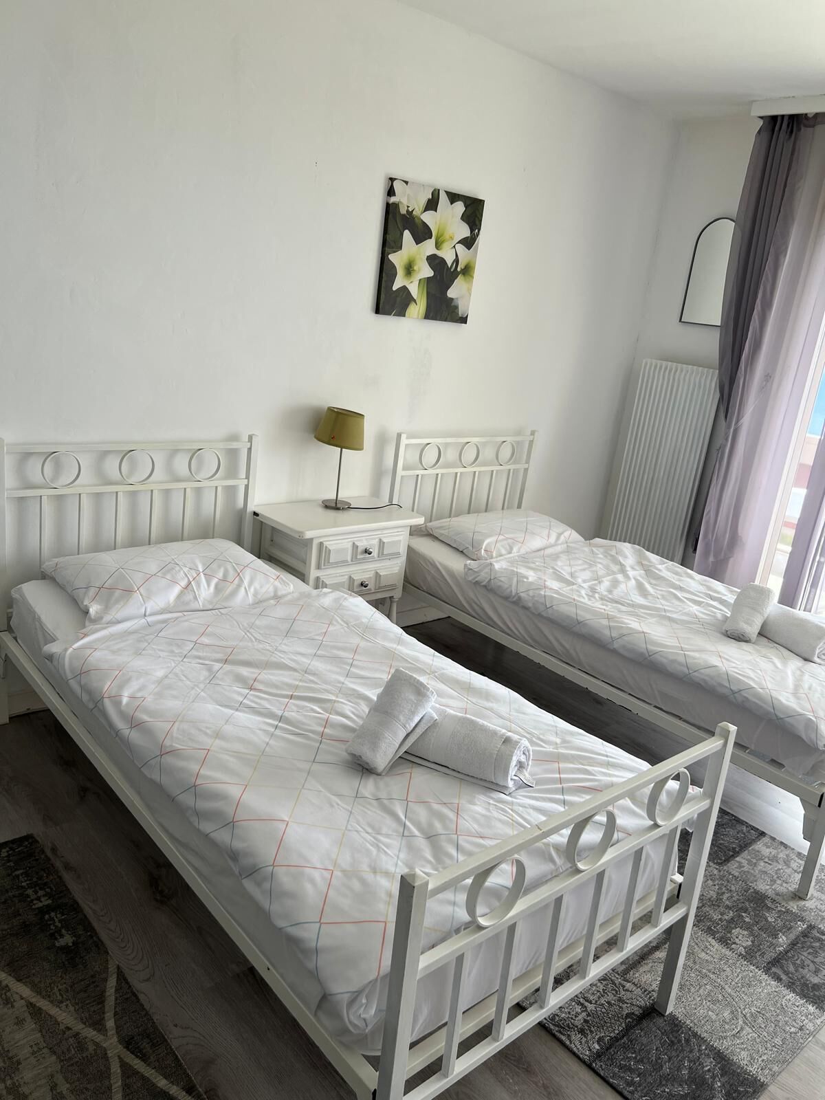 A hotel room with two single beds, a bedside table with a lamp, a radiator, and a window with curtains.