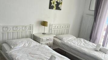 A hotel room with two single beds, a bedside table with a lamp, a radiator, and a window with curtains.