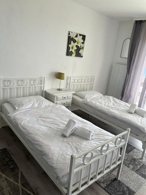 A hotel room with two single beds, a bedside table with a lamp, a radiator, and a window with curtains.