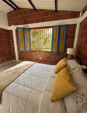 A room with a bed, a window with bars, a lamp, and a brick wall.