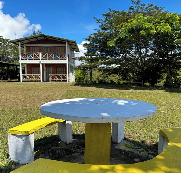 A two-story building with a balcony, a round table with benches, and a grassy area with trees.