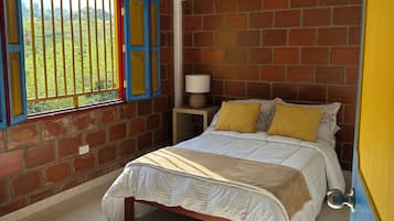 Ein Schlafzimmer mit Ziegelwand, einem Bett mit weißer und beiger Bettdecke, einem Nachttisch mit Lampe und einem Fenster mit Blick auf Grünflächen.