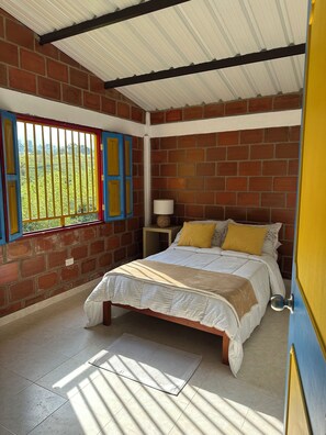 A bedroom with a brick wall, a bed with a white and beige bedspread, a nightstand with a lamp, and a window with a view of greenery. - A cabin nestled among the mountains, birds, and a magical view in El Queremal (Valle del Cauca)