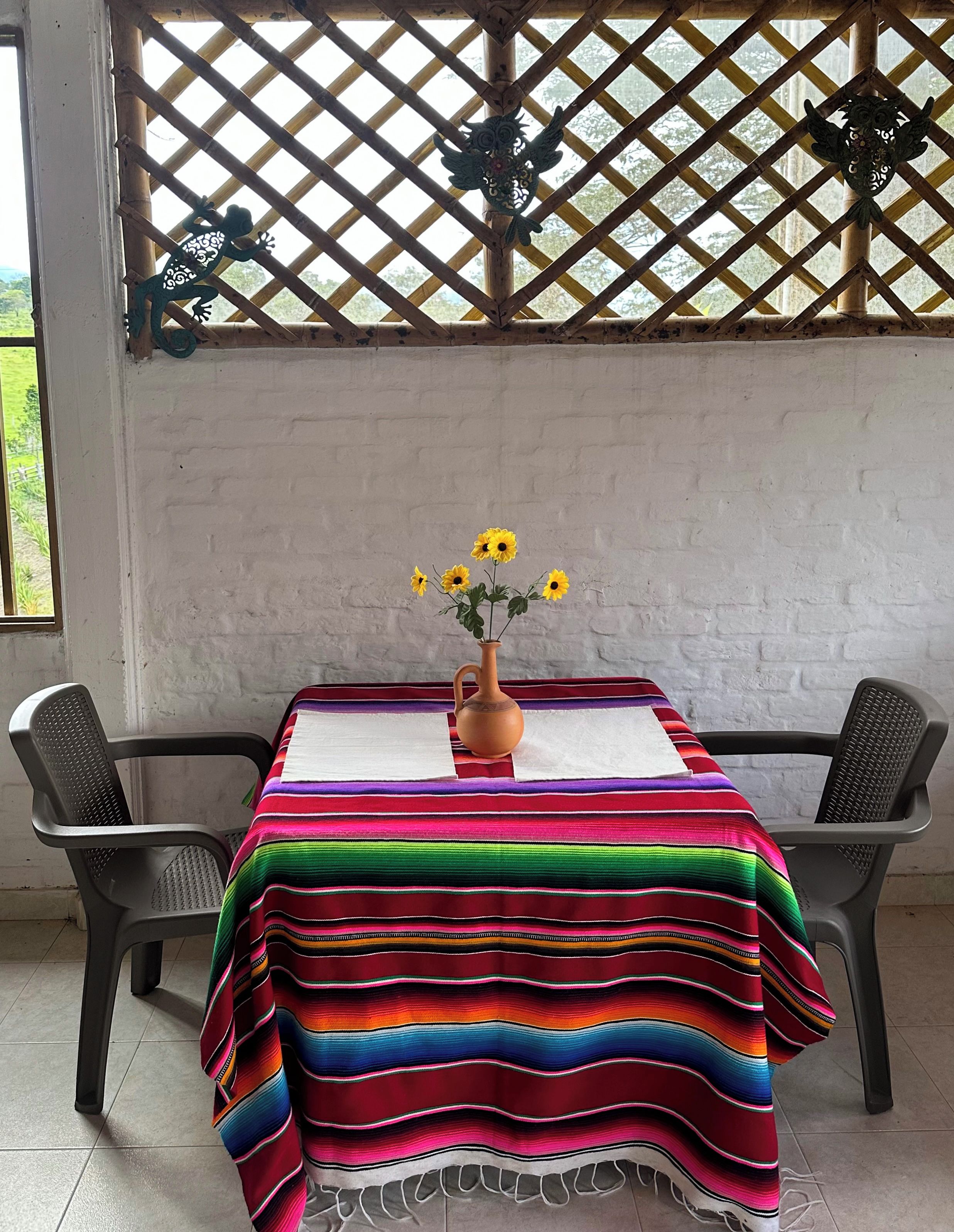 Une table recouverte d’une nappe rayée aux couleurs vives, deux chaises et un vase rempli de fleurs jaunes.