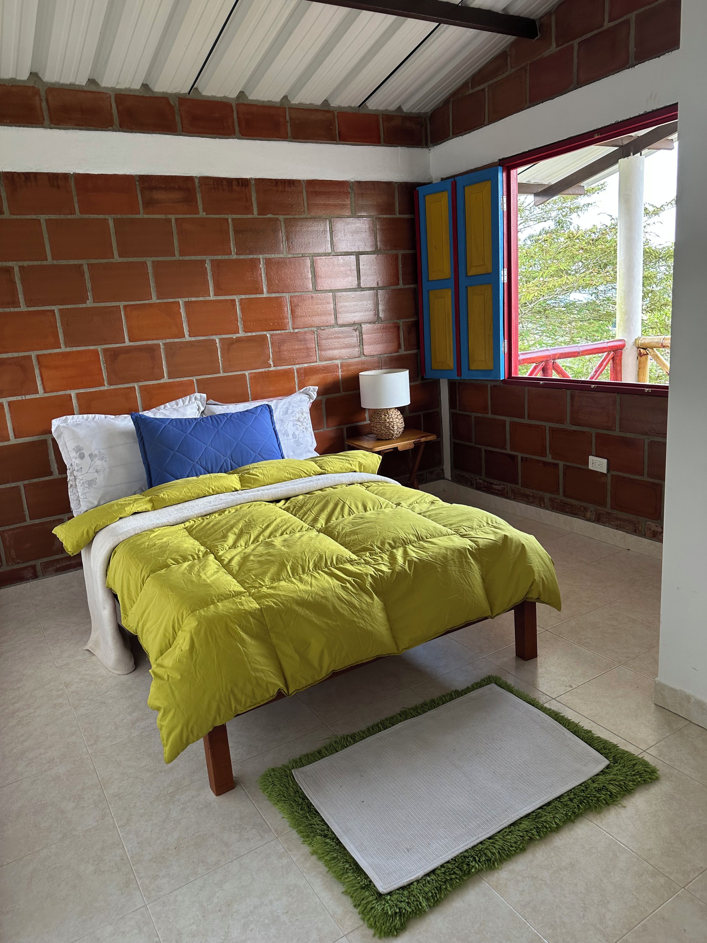 A bedroom with a brick wall, a bed with a yellow bedspread, a nightstand with a lamp, and a window with red railings.