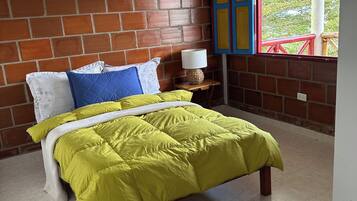A bedroom with a brick wall, a bed with a yellow bedspread, a nightstand with a lamp, and a window with red railings.