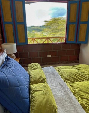 Una habitació amb un llit, una finestra que dona a vegetació i una paret de maó.