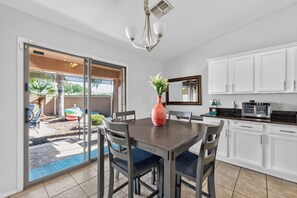 Une cuisine moderne avec des meubles blancs, une table à manger avec des chaises, un vase rouge et une porte-fenêtre coulissante donnant sur un espace extérieur.