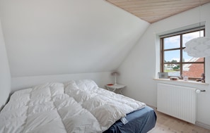 Et soveværelse med skrå vægge, en seng med hvide sengetøj, et lille natbord med en lampe, et vindue med udsigt til en murstensbygning, og en radiator.