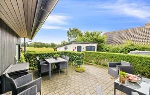 En udendørs terrasse med fletmøbler og et lille bord, en hvid bygning med blå døre, og en grøn hæk i baggrunden.