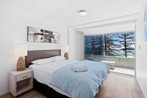 A modern bedroom with a large bed, bedside lamps, a view of trees through a glass door, and a ceiling light. - Goodwin Towers with Beach Views! (Gold Coast)