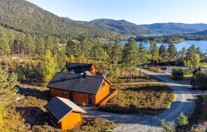 Une cabane dotée d’un toit en pente, entourée d’une zone boisée, avec un lac et des montagnes en arrière-plan.