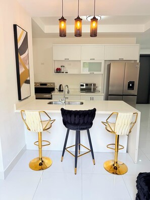 A modern kitchen with white cabinetry, a central island with bar stools, a stainless steel refrigerator, and pendant lighting. - The Uptown Apartment 
Kingston’s Luxurious apartment perfect for up to 3 persons (Kingston)