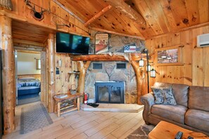 A log cabin interior with a stone fireplace, a television mounted on the wall, and a leather sofa. - Year-Round Trail Access | Cozy Cabin on Lake (Cable)