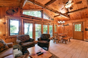 A cabin interior with wooden walls, large windows, a dining area, and a fireplace. - Year-Round Trail Access | Cozy Cabin on Lake (Cable)