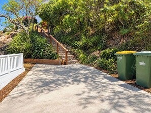 A concrete pathway leading to a wooden staircase surrounded by lush greenery and two green dumpsters. - Hillside Vista| West End Retreat w/Panoramic Views (West End)