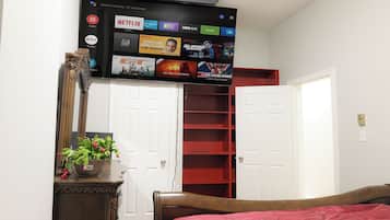 A bedroom with a bed, a dresser, a mirror, a potted plant, and a TV mounted above a door displaying various streaming services.