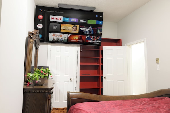 Ein Schlafzimmer mit einem Bett, einer Kommode, einem Spiegel, einem Topfpflanzen und einem Fernseher, der über einer Tür angebracht ist und verschiedene Streaming-Dienste anzeigt.