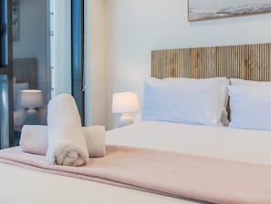 Ein Hotelzimmer mit einem Bett, Kissen, einem Kopfteil, einer Nachttischlampe und einem aufgerollten Handtuch auf dem Bett.