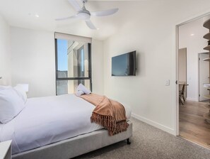A modern bedroom with a bed, a ceiling fan, a large window, a flat-screen TV, and a view of the outside. - Rosa | Hemingway Palm Beach | 3 Bedroom Ocean (Gold Coast)