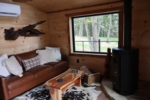 Ein Interieur einer Hütte mit einem braunen Ledersofa, einem Holztisch, einem schwarzen Kaminofen und einem Fenster mit Blick auf Bäume.