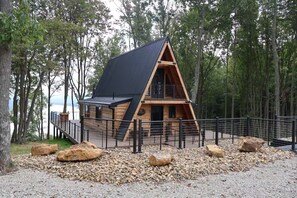 Eine Holzhütte mit geneigtem Dach, umgeben von einer Schotterfläche und einem Metallzaun, inmitten eines bewaldeten Geländes.