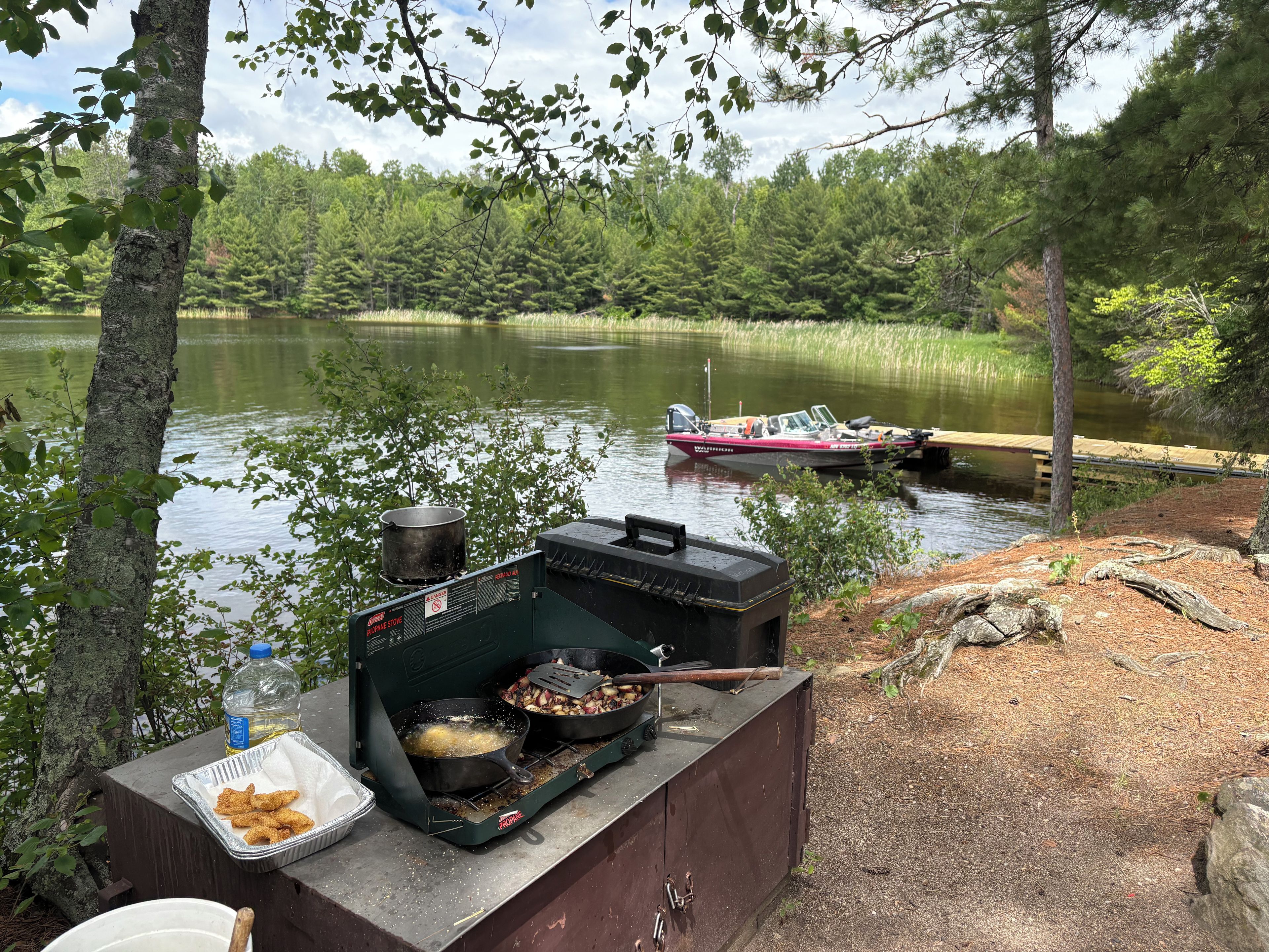 A portable grill with food, a pot, and a bottle on a wooden platform by a lake with a boat in the background.