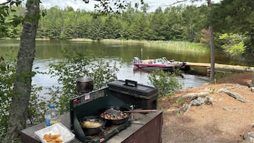 A portable grill with food, a pot, and a bottle on a wooden platform by a lake with a boat in the background.