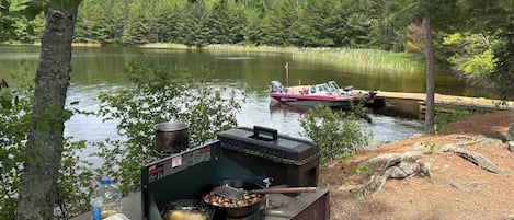 A portable grill with food, a pot, and a bottle on a wooden platform by a lake with a boat in the background.