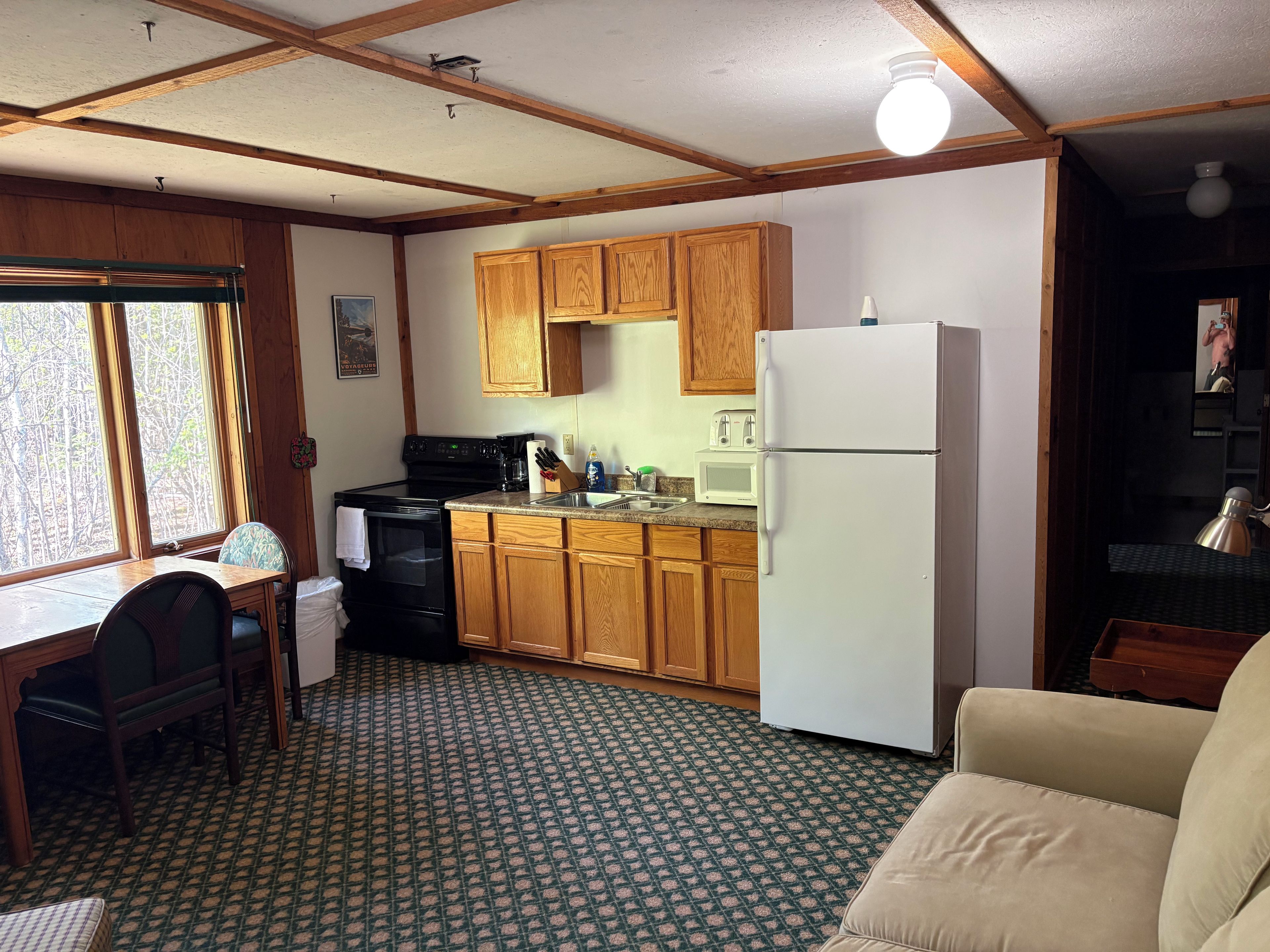 A compact kitchen with wooden cabinets, a white refrigerator, a microwave, and a stove. There is a dining table with chairs and a patterned carpet.
