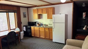 A compact kitchen with wooden cabinets, a white refrigerator, a microwave, and a stove. There is a dining table with chairs and a patterned carpet.