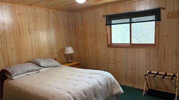 A wooden-paneled room with a bed, a nightstand, a lamp, a window with blinds, and a shoe rack.