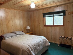 Een kamer met houten lambrisering, een bed, een nachtkastje, een lamp, een raam met jaloezieën en een schoenenrek.