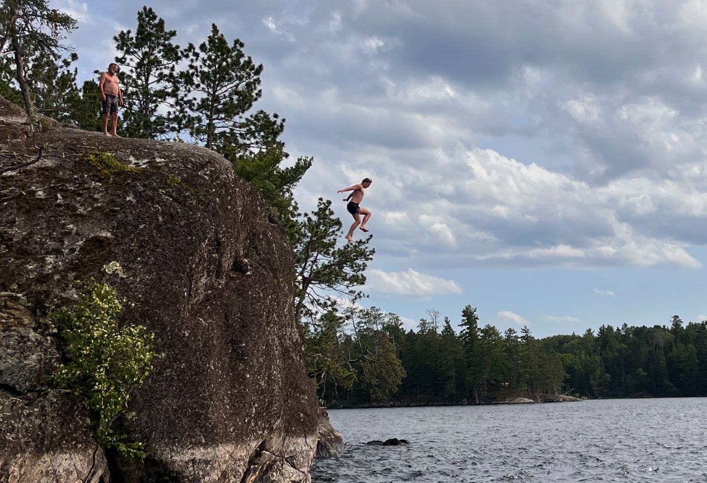 A person jumping off a rocky cliff into a body of water, surrounded by trees and a cloudy sky.