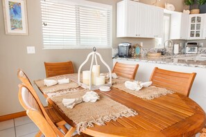 A wooden dining table with a woven placemat, surrounded by wooden chairs, set in a kitchen with white cabinets and a window with blinds. - Hooked on Little Torch: 3BR Escape with 40' Dock, 15,000 lb Boat Lift & Elevator (Little Torch Key)
