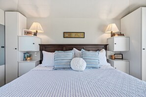 A neatly arranged bedroom with a bed, two bedside tables, a nightstand, a wall-mounted shelf, and a framed picture above the bed. - The Winthrop - Beachfront Cape Cottage with private beach (Barnstable)