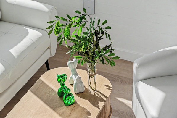A room with a white sofa, a round wooden coffee table, a glass vase with green plants, and two white ceramic figurines.