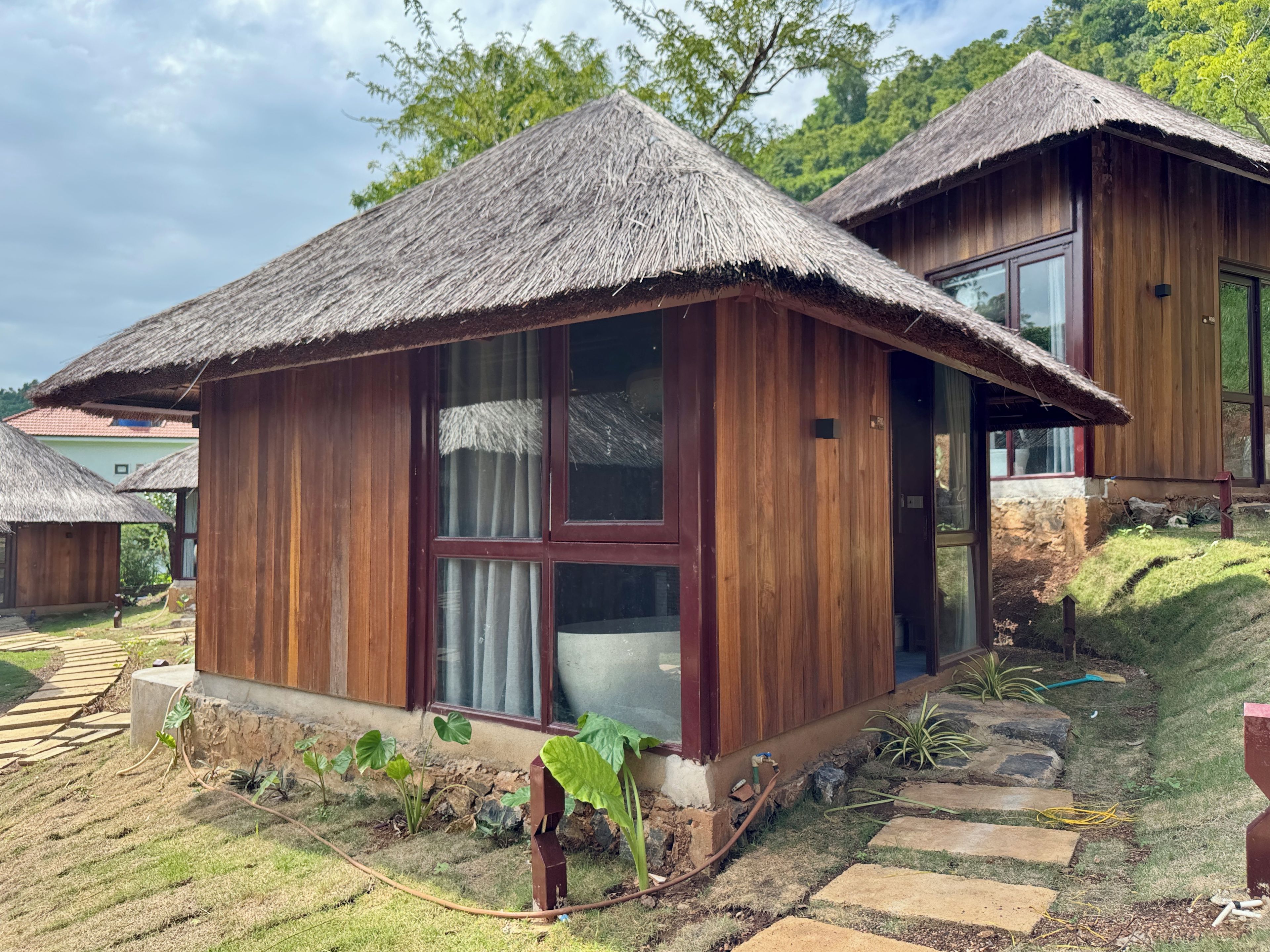 Une maison en bois avec un toit de chaume, une grande fenêtre et un petit jardin agrémenté de pierres plates servant de pas.