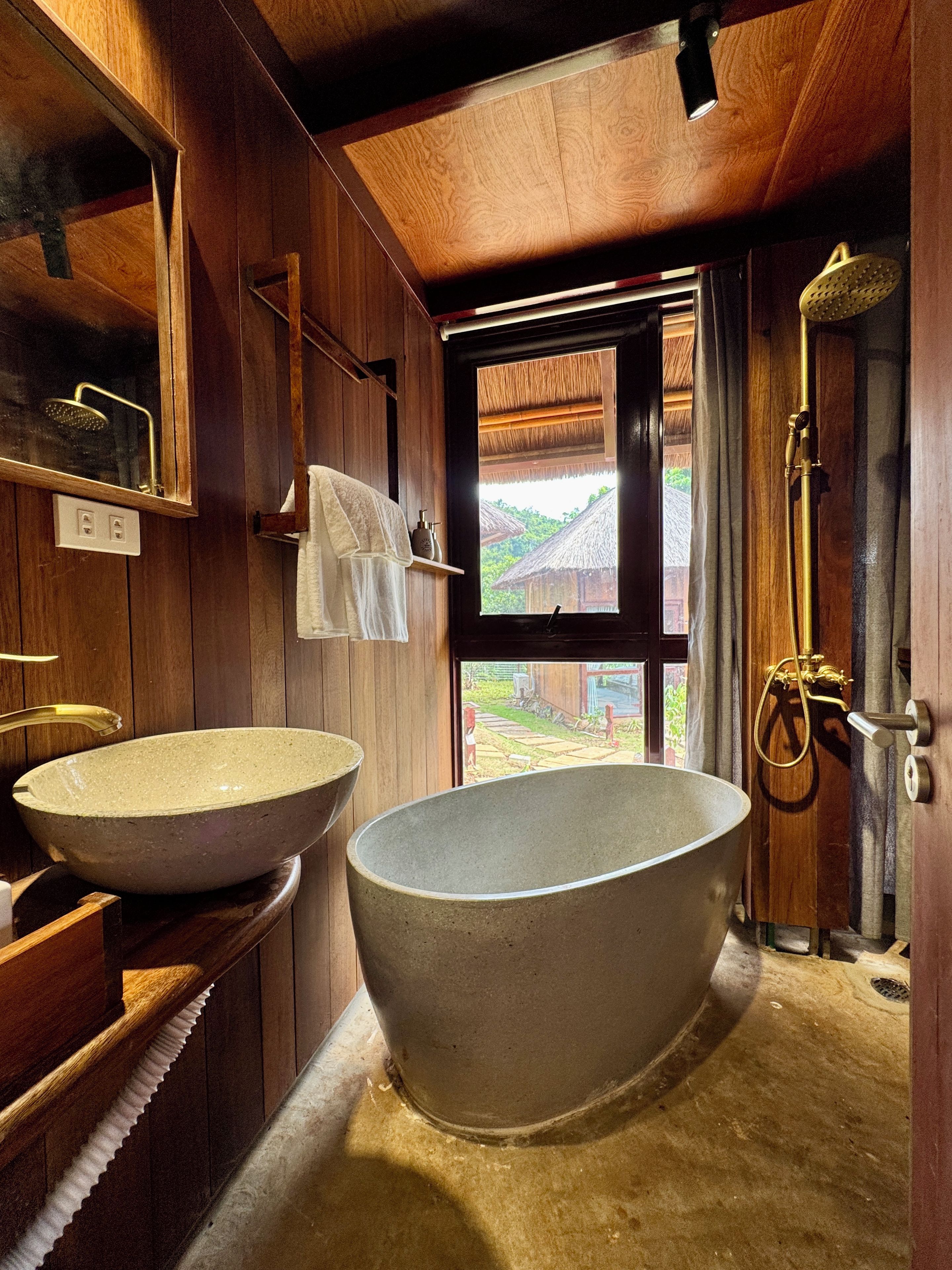 Une salle de bain avec deux grands lavabos en pierre, des murs en bois, une douche et une vue sur un bâtiment à toit de chaume à l’extérieur.