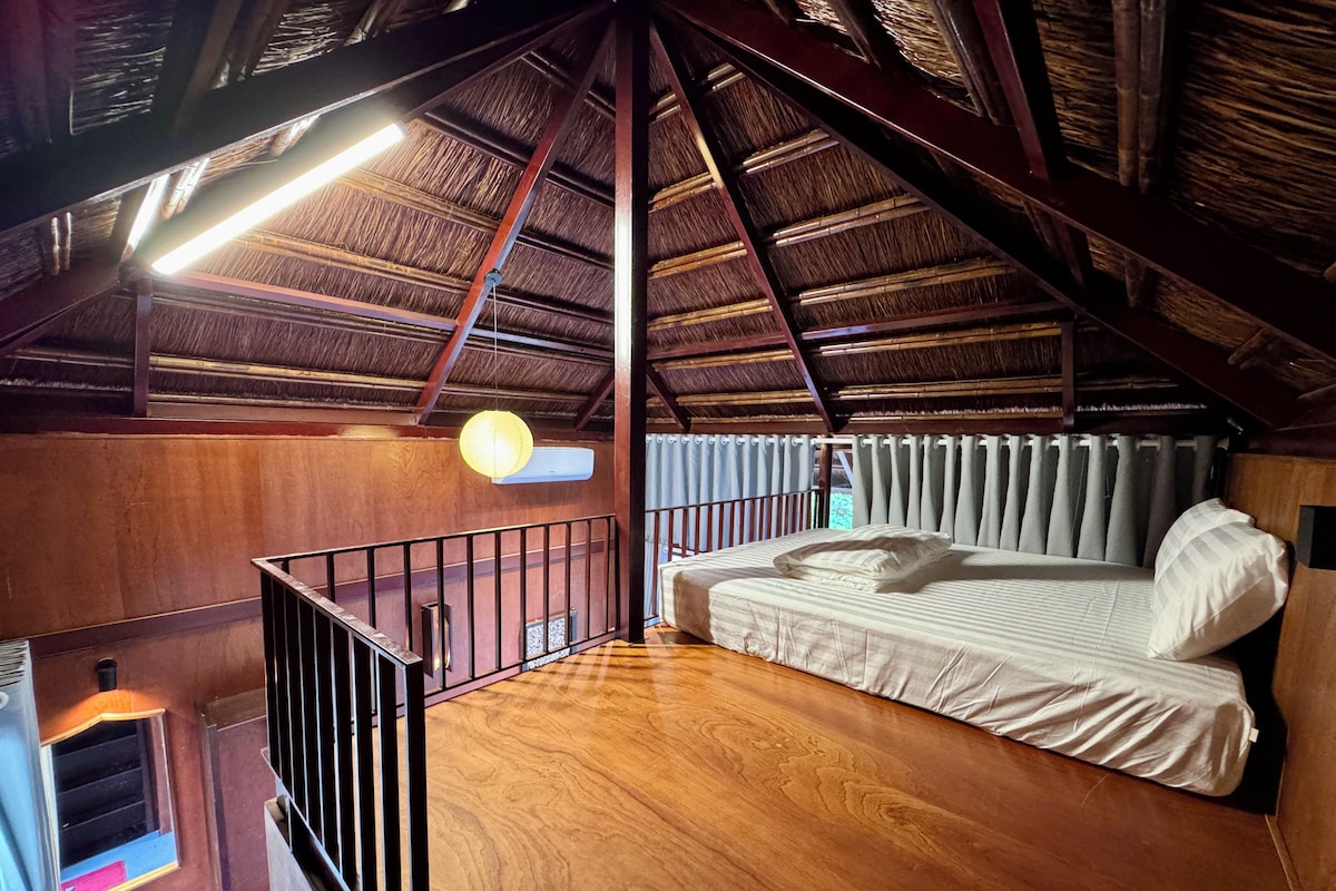 Een kamer met een houten plafond, een bed met witte lakens, een klein raam en een luchtverkoeler aan de muur.