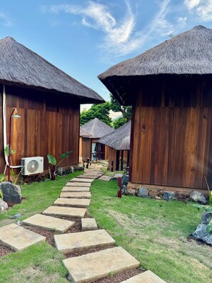 A pathway leading to wooden buildings with thatched roofs, surrounded by greenery.