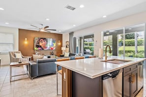 A modern kitchen with a central island, a dining area with a table and chairs, a seating area with sofas, a television mounted on a wooden wall, and a large window offering a view of the outdoors. - Experience Magic and Luxury Just Minutes from Disney (Florida)