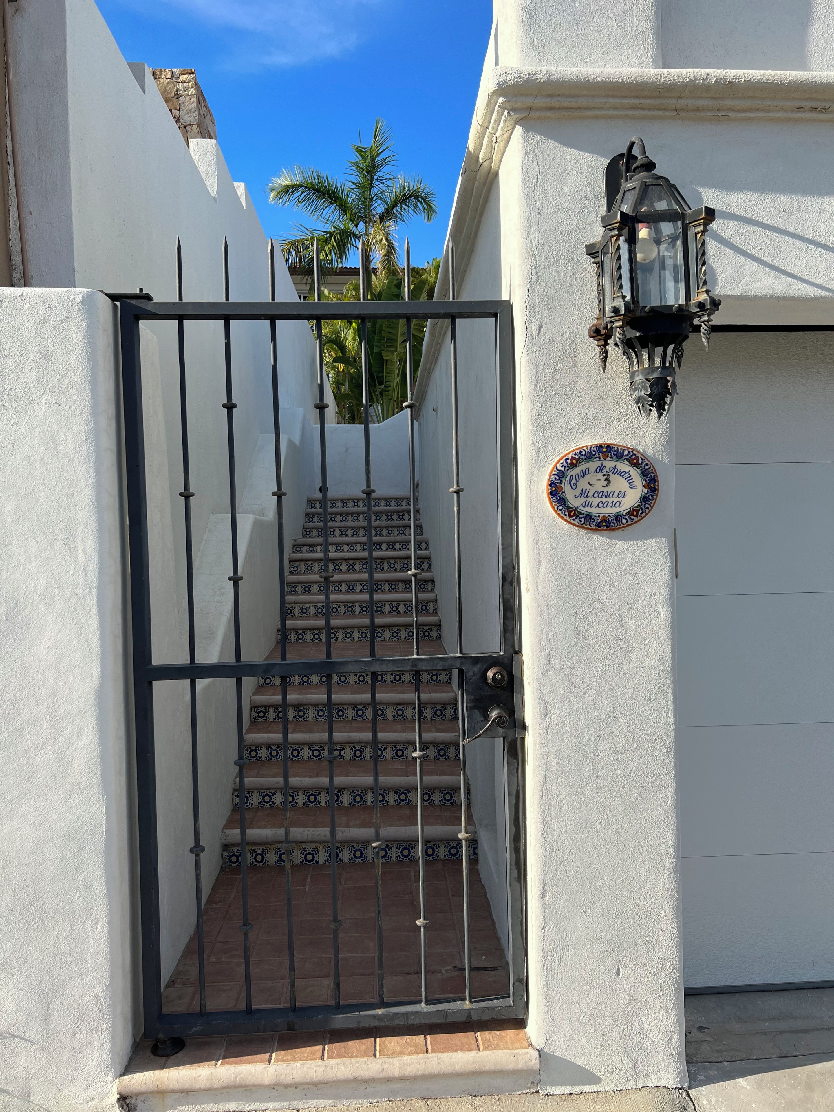 Une entrée fermée par un portail, avec un escalier carrelé menant à un bâtiment blanc, une lanterne décorative et un panneau portant un message.